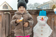 © Katharina - Happy child smiling at camera holding snack next to funny snowman on wooden balcony outdoor winter activity portrait joy authentic emotion child care seasonal promotion