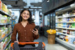© Liubomir - Young indian woman smiling while pushing a shopping cart and using a smartphone to browse products and compare prices in a modern grocery store aisle, candid and cheerful