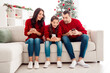 © deagreez - Family in red sweaters excitedly looking at smartphones together on a sofa by a decorated Christmas tree, sharing holiday moments and laughter with their daughter between parents