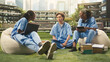 © DC Studio - Medical students discussing coursework with laptop and study materials, stacked books and notes while seated on beanbags in sunny modern campus courtyard.
