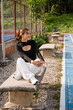 © VICTOR A ARGUELLO - Young latina woman sitting on a stone bench in a park, enjoying leisure time outdoors and looking away, dressed in fashionable casual attire during daylight