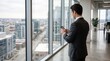 © Jun - Businessman standing by window looking at smartphone, cityscape view from high rise office building, corporate executive checking mobile device while overlooking urban skyline