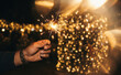 © Nina - Man holding a burning sparkler during a festive celebration at night with beautiful golden bokeh lights background representing holiday joy
