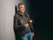 © faisal - A mature caucasian male leans casually against a textured concrete wall holding a bright lit sparkler. He wears a rugged brown leather jacket over a grey hoodie, gazing away into the