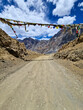 © Travel 'n' Lifestyle - View of the rugged Penzi La road in Zanskar, Ladakh, India, featuring colorful prayer flags strung across the dirt path against a backdrop of dramatic mountain peaks and a blue sky.