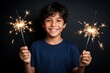 © Maryna - Happy Brazilian boy holds lit sparklers in both hands. Young kid smiles brightly against dark background. Child celebrates festive occasion with glowing fireworks. Joyful kid enjoys magic light,