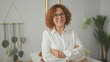 © Krakenimages.com - Hispanic woman with glasses and arms crossed in studio setting, smiling amid macrame wall hanging, bed and plants; confidence presence.