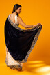 © Wavebreak Media - Adult Indian woman posing, lifting navy velvet pallu with gold border in studio on orange backdrop