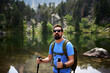 © CarlosBarquero - A Caucasian man stands with trekking poles, sunglasses and a green backpack beside a forested lake. Photographed in the Aiguestortes area of the Spanish Pyrenees.