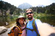 © CarlosBarquero - A Latina African American woman and a Caucasian man smile into the lens beside a mountain lake, with a yellow backpack and green backpack visible. Photographed in the Aiguestortes area of the Spanish