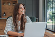 © Daniel - Woman studies online on a laptop at a wooden table, typing while looking away with a thoughtful smile. Home learning, coursework and education fit a bright, quiet study space with natural daylight.