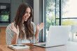 © Daniel - Woman greets colleagues during a laptop video call at a wooden table. Her enthusiastic wave and smile support remote work, workplace communication, online meetings and a welcoming team connection.