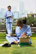 © DC Studio - Medical student in blue scrubs writes notes beside an open textbook and stacked books on a sunny campus lawn while a classmate studies nearby outdoors.