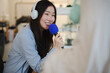 © Studio Marmellata - A young Asian woman wearing white headphones smiles happily while speaking into a blue microphone during a podcast recording session in a bright modern studio.