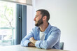 © simona - Young man with a beard and blue shirt looking out a bright window from indoors, sitting at a wooden table with arms crossed, contemplating and reflecting on his thoughts