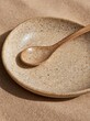 © Elena - Close-up of a small, round plate with a wooden spoon resting on it. the plate is made of a light-colored material with a speckled texture, and the spoon has a smooth, polished finish.