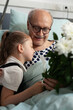 © DC Studio - Affection, hug and hospital visit with a girl leaning close to a smiling senior patient beside white flowers, a tender bedside moment for family support, comfort, love and emotional connection.