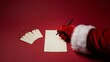 © MSTMAYA - Santa claus hand in red glove writing on a blank white paper sheet with blank gift tags on a red background, for christmas holiday, gift preparation
