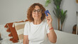 © Krakenimages.com - Woman holding creditcard to temple while seated on bed in a bedroom with decorative pillows, plant and neutral wall behind her; financial frustration.