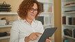 © Krakenimages.com - Woman taps tablet with both hands in a building office lined with bookshelves and binders, wearing glasses and a white shirt; focus productivity.