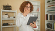 © Krakenimages.com - Woman middle aged with curly hair holding a tablet, hand to forehead while reading a screen in a building with bookshelves and files; frustration focus.