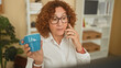 © Krakenimages.com - Woman holding smartphone to ear and blue mug in hand, wearing glasses and curly hair while sitting at desk with computer in a building; concentration routine.