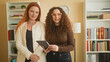 © Krakenimages.com - Two women colleagues hold a tablet together, one tapping the screen while both smile, bookshelves and a clock visible in an office room inside a building; friendly collaboration trust.