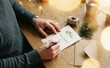 © Nina - Adult man writing a Christmas greeting card at wooden table with festive decoration and envelope for winter holiday season