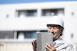 © kapinon - 青空の下でマンションを背景にタブレットを使って施工管理やマンション管理業務、大規模修繕を行うヘルメットと作業着姿の中年男性