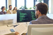 © Studio Romantic - Developer programmer coding software on computer monitor. The man coder in modern office reviews documents and code as colleagues discuss tasks nearby. Concept software development and teamwork.