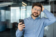 © Tetiana - A close-up portrait of a young, upset man indoors; he is holding a phone in one hand, clutching his head with the other, and looking at the camera with a disappointed expression