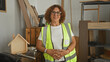 © Krakenimages.com - Woman carpenter in a yellow reflective vest stands with hands clasped beside wooden house model in building; confidence craftsmanship.
