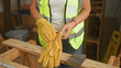 © Krakenimages.com - Woman in reflective vest putting on leather glove with bare hands over wooden plank on sawhorse inside building; focused safety.
