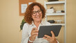 © Krakenimages.com - Woman holding a tablet and tapping the screen with a stylus in building, smiling while seated in front of shelf and plant; contentment.