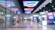 © Fauzan - Blurred indoor background of a modern commercial exhibition hall, featuring glowing geometric ceiling lights, colorful illuminated display booths, and a spacious convention center walkway.