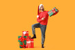 © Pixel-Shot - Christmas shopping and holiday sales, happy man in santa hat holding gift box with pile of presents on yellow background