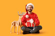 © Pixel-Shot - Christmas gift giving and holiday celebration, happy man in Santa hat holding present with wooden reindeer on yellow background