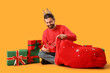 © Pixel-Shot - Christmas gift giving and holiday celebration, man wearing reindeer antlers putting presents into a large red sack on a yellow background