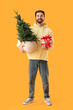 © Pixel-Shot - Christmas shopping and holiday preparation, happy man holding a small pine tree and gift boxes on a yellow background