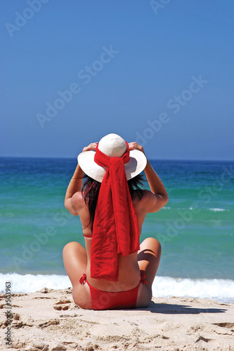 girl sitting on sandy beach in the sun adjusting hat. blue sky,