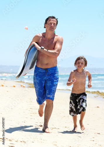 young father and son running along beach with surfboard