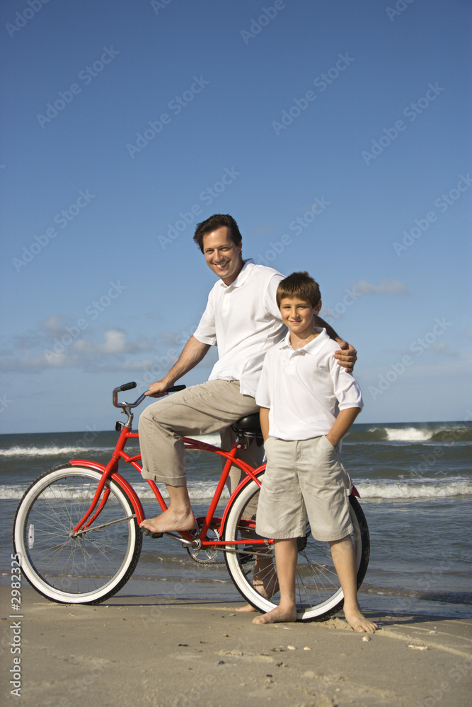 Father on bike with arm around son on beach.