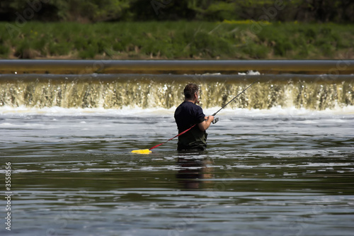 Man wading in a river and fly fishing with a low waterfall 