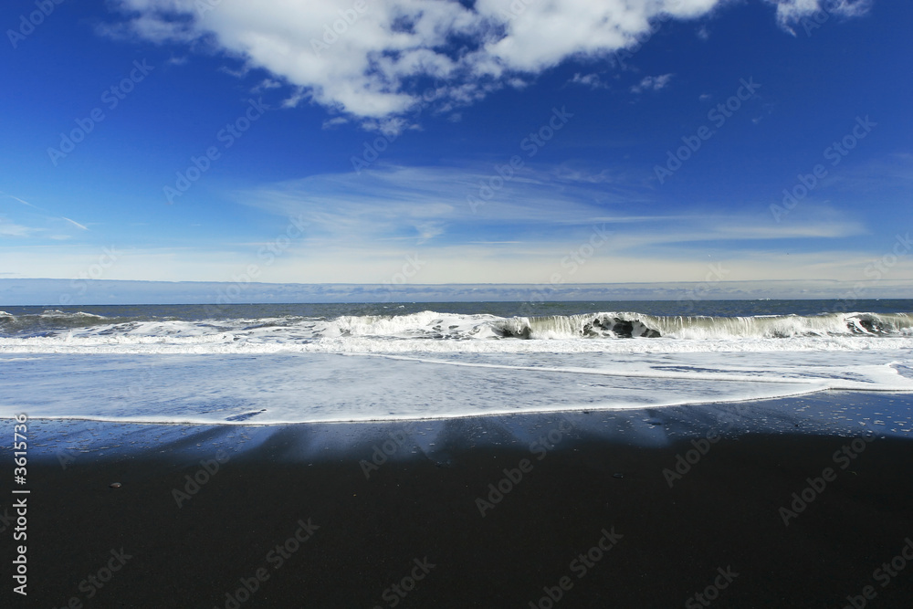 waves crashing on a sandy black beach , deep blue sky