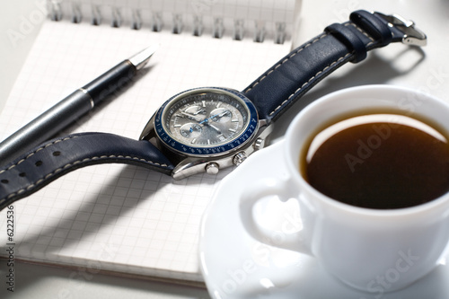 Watch and pen over notebook with cup of coffee near by