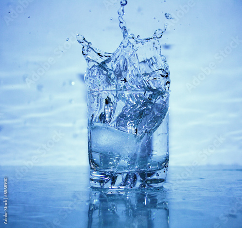 Glass of splashing water with ice on blue background