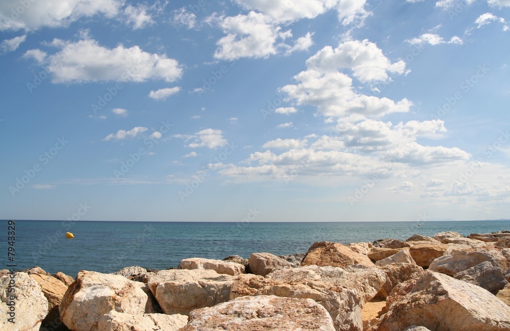 Blue sky with clouds above blue sea and stones in front