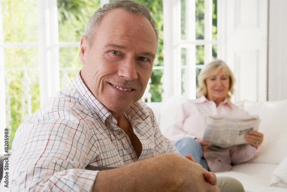 Man in living room smiling with woman in background reading news Man in living room smiling with woman in background reading news