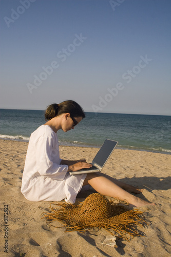 Woman sitting on beach in white dress working on laptop