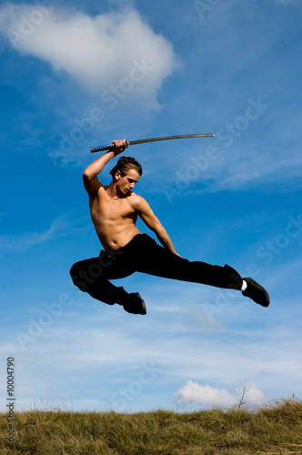 Young man with samurai sword jumping against blue sky.